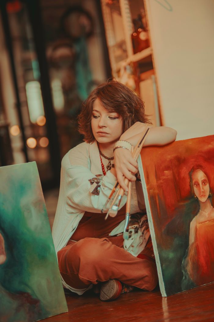 A female artist sits thoughtfully in a studio, surrounded by her expressive portraits.